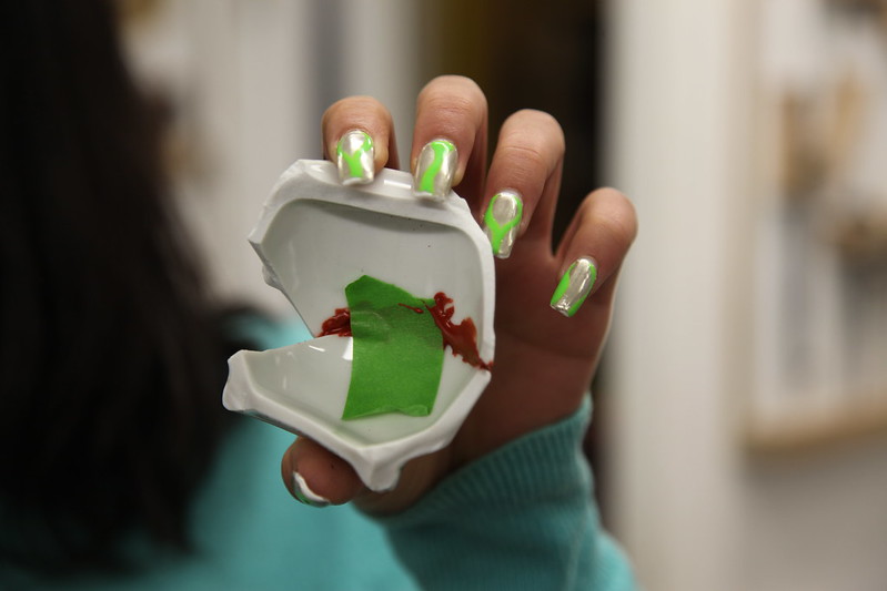 Repurpose rather than repair - a person with green and silver nail art holds up fragments of ceramics that they have stuck together with red resin and green tape, creating a bold artistic repair rather than an invisible repair